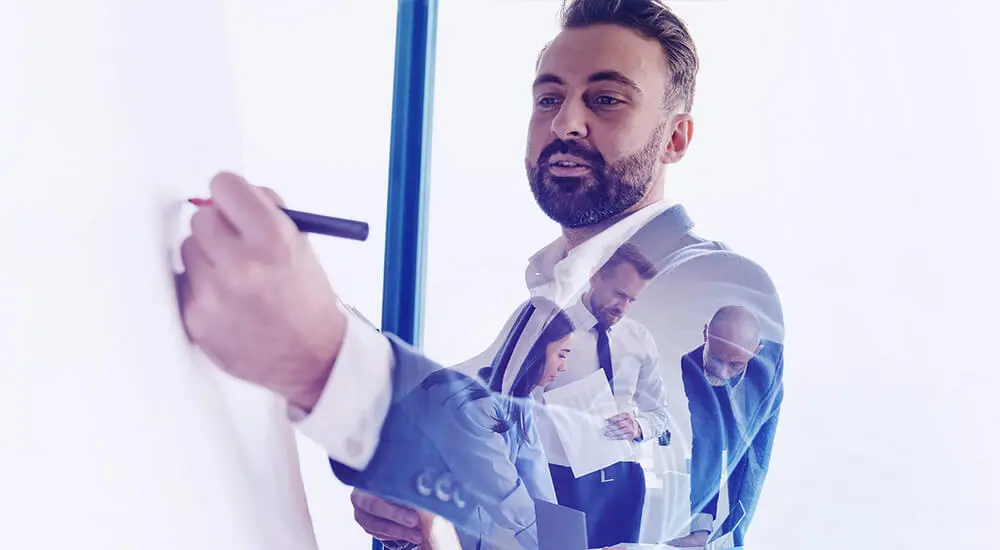 Photogrsph of man writing on whiteboard with meeting in background